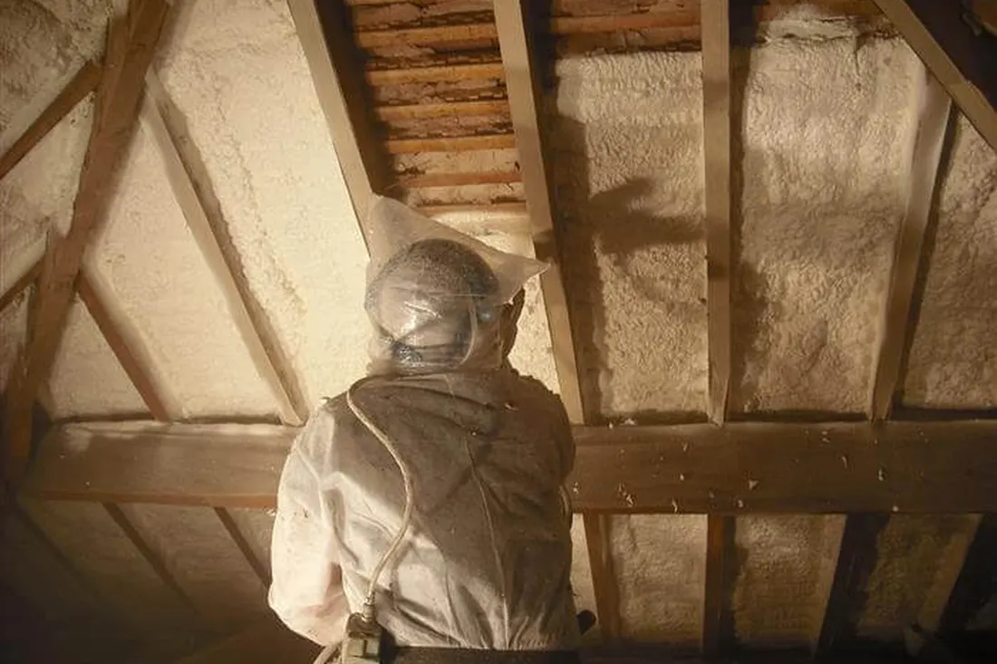 Worker in a hood and suit applying spray foam between attic rafters