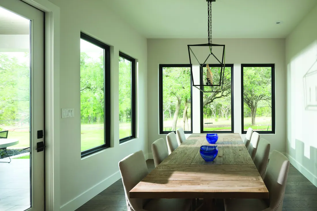 Dining room with black-framed picture windows overlooking green trees and a rustic wood table