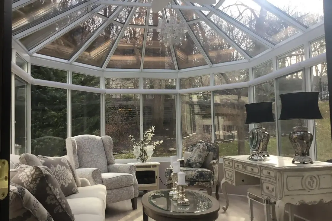 White-framed glass sunroom conservatory furnished with upholstered seating and a desk overlooking trees