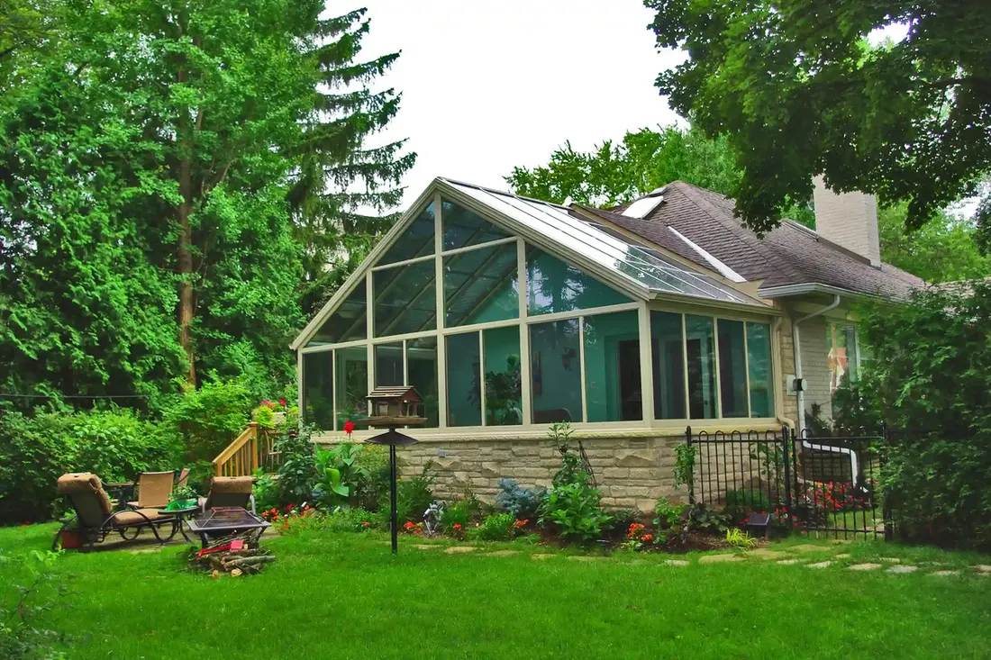 Green-framed glass sunroom addition behind a stone house and lawn