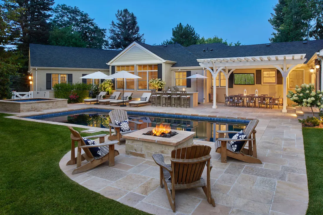 Backyard swimming pool surrounded by stone patio, fire pit, wooden chairs, and white pergola