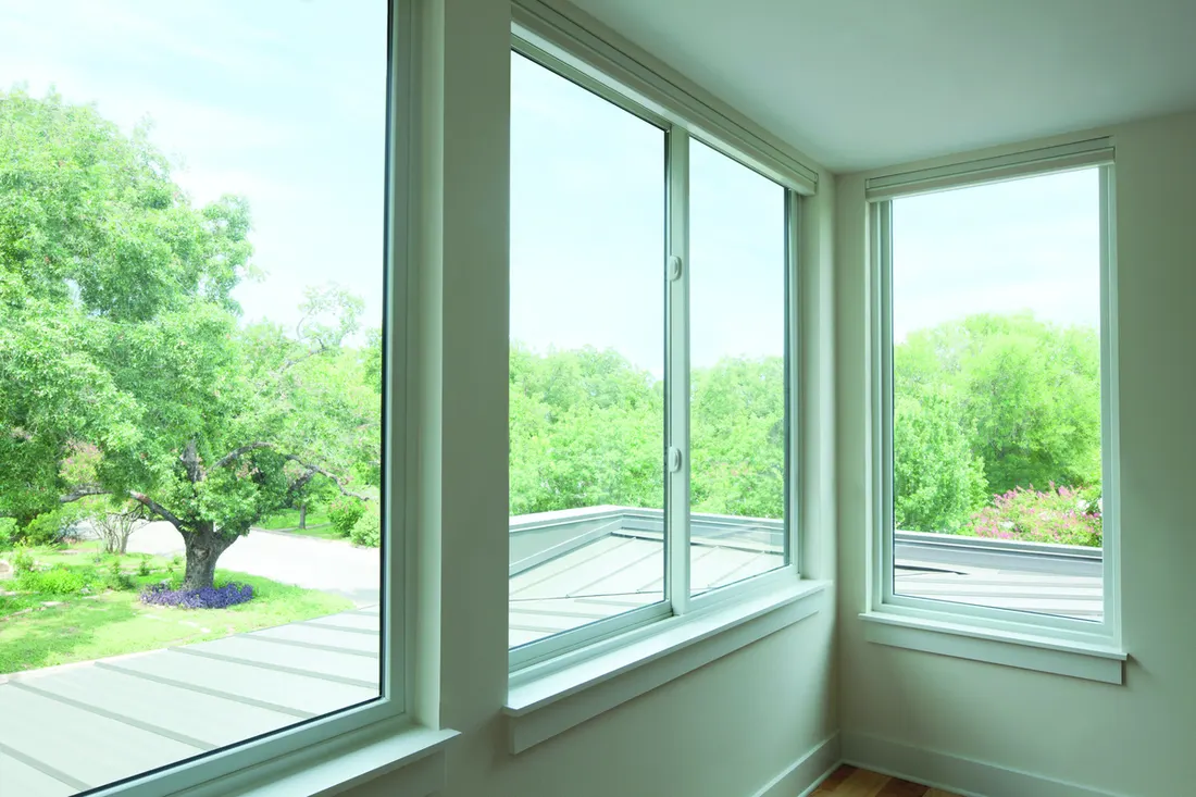 Large white frame windows in a sunlit room overlooking green trees and outdoor rooftops