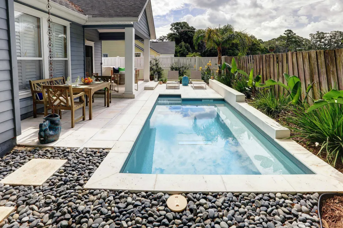 Rectangular plunge pool surrounded by light stone pavers, dark river rocks, and a backyard patio area