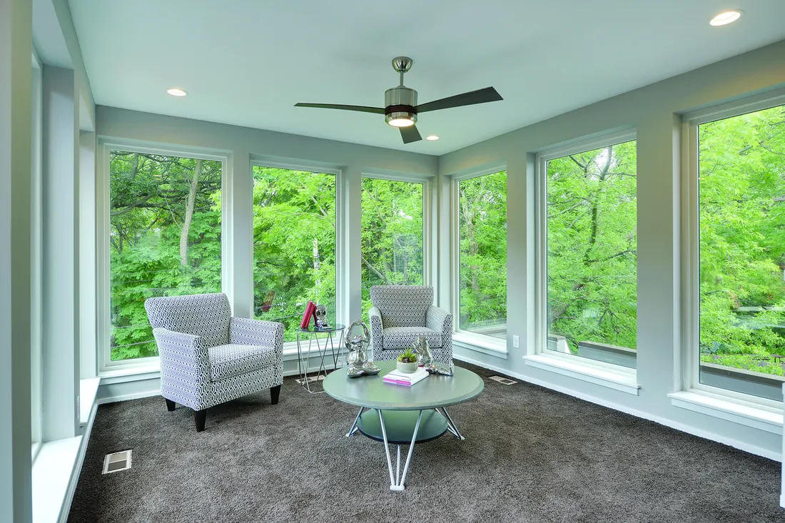 Sunroom lined with large white-framed picture windows overlooking bright green outdoor trees