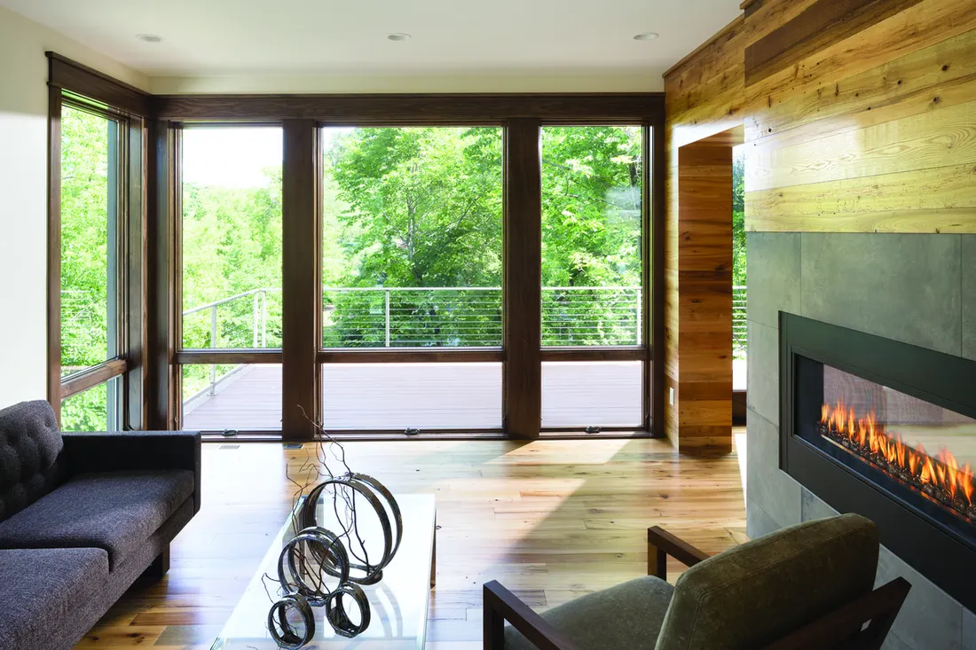 Dark wood-framed picture windows in a living room overlooking an outdoor deck and green trees