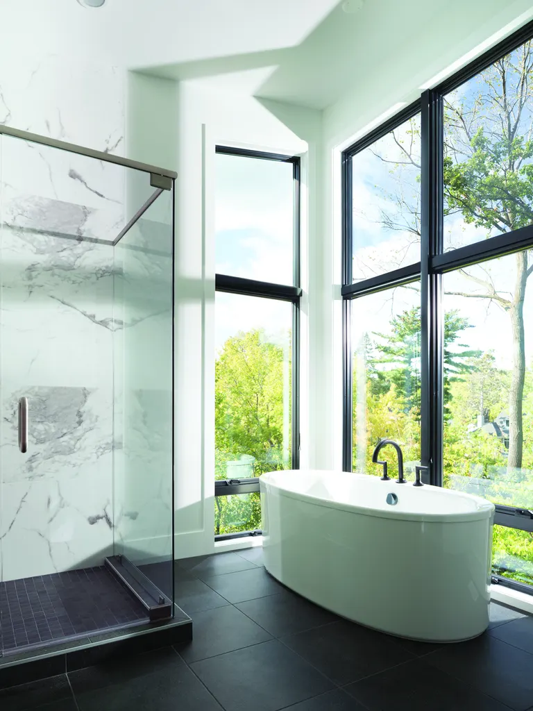 Large black-framed windows overlooking lush green trees beside a white freestanding soaking tub